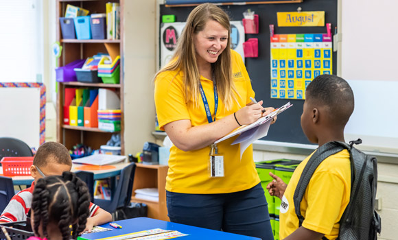 Teacher wearing Moss Street Partnership School yellow shirt smiling at student.