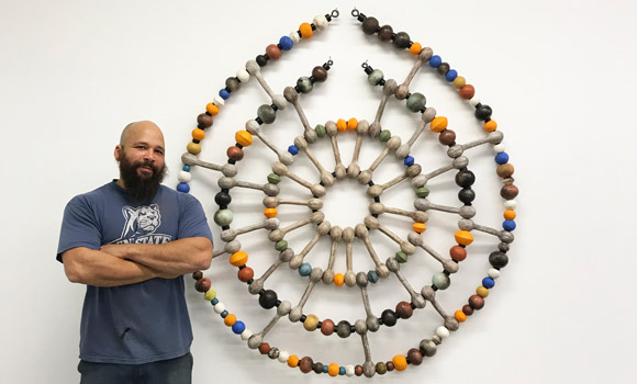Alumnus and artist Sharif Bey standing beside his artwork, circular with earthen colors.