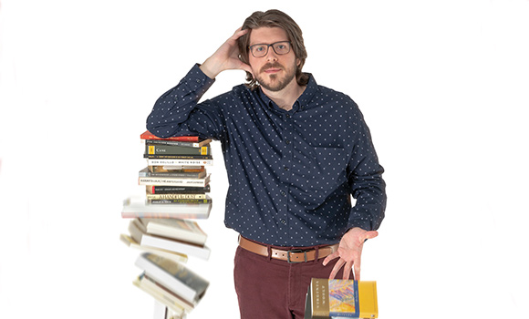 Photo of Matt Phillips leaning on a pile of books that is falling