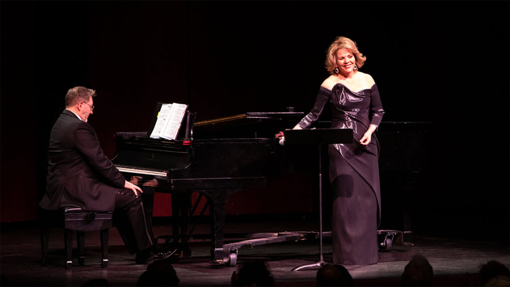 Renee Fleming smiles on stage next to piano accompaniment.