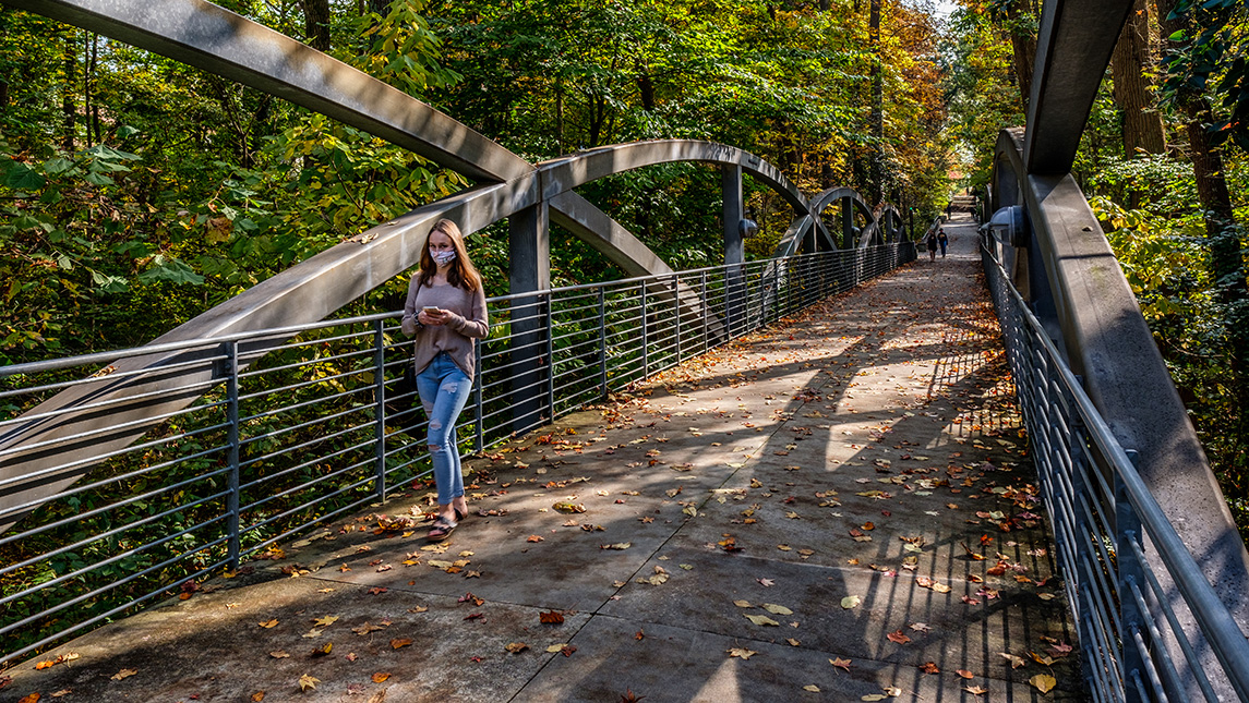 student walking on campus bridge with fall leaves