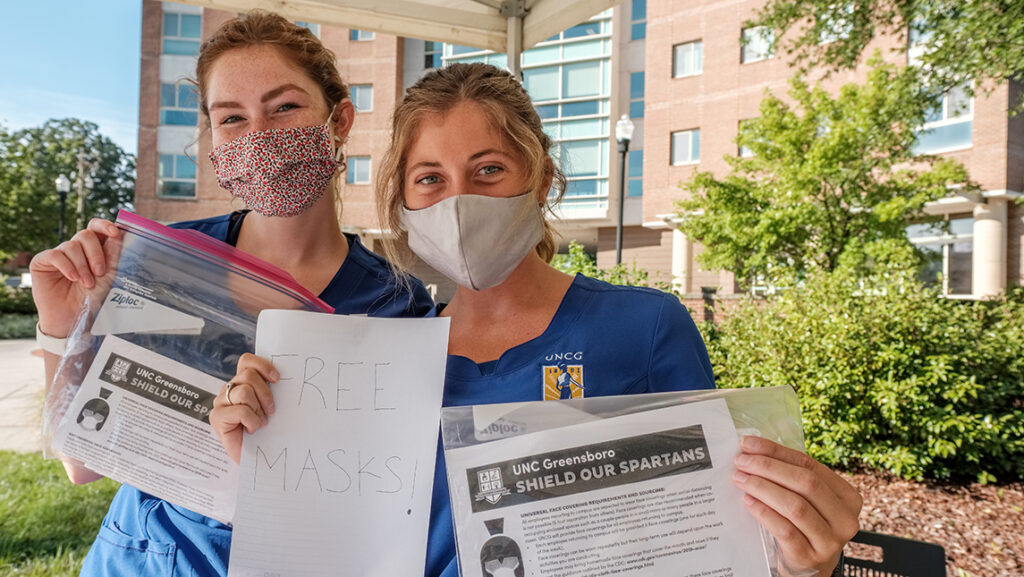 Two nursing students wearing face coverings pose with bags of free masks to be given out to campus community