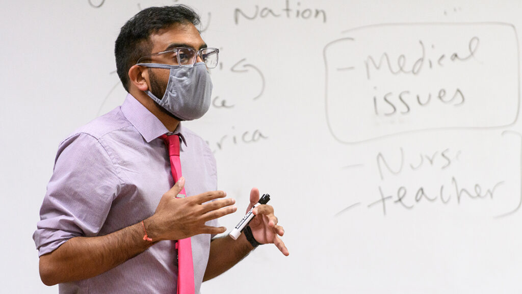 Professor wearing face covering teaches in front of a white board