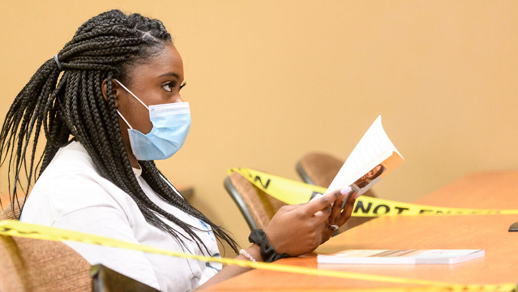 Student wearing face covering peers over a piece of literature in class with caution tape on either side of her seat