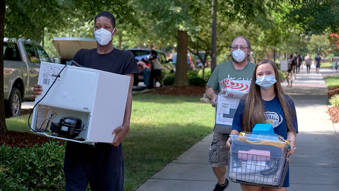 Three people wearing masks and carrying belongings into the dorm during move-in