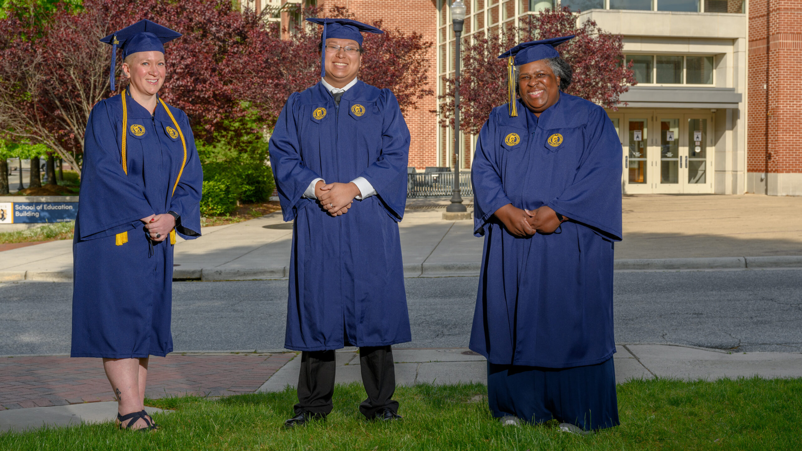 Jessica Jeffus, Jimmy Vang, and Lynnette Pitts pose in their cap and gown in front of the School of Education Building