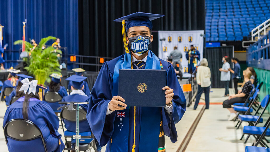 Student holding up diploma cover with UNCG branded mask at commencement