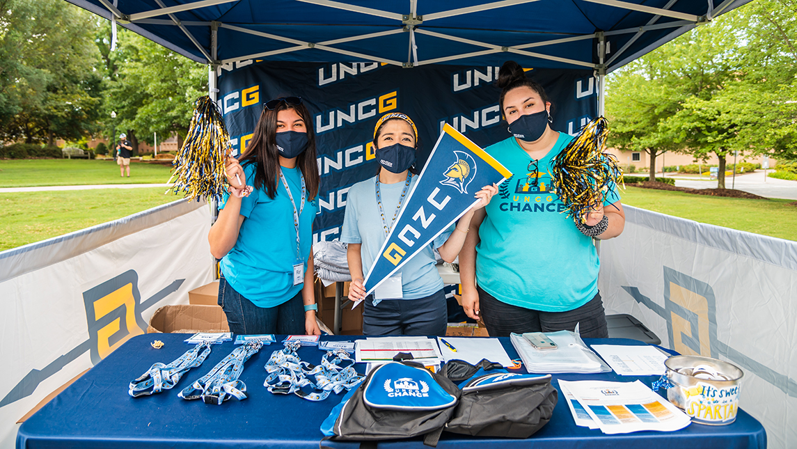 Three students pose in masks  with UNCG pom-poms and banner under a blue tent with CHANCE swag