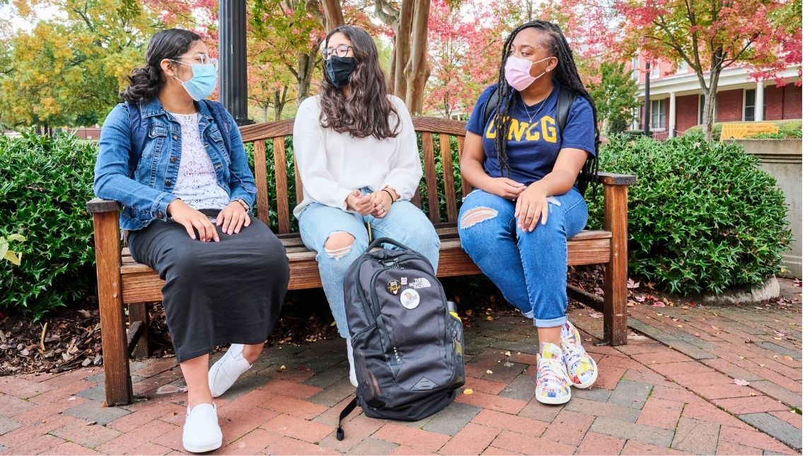 Three students on a bench talking and wearing face coverings surrounded by trees