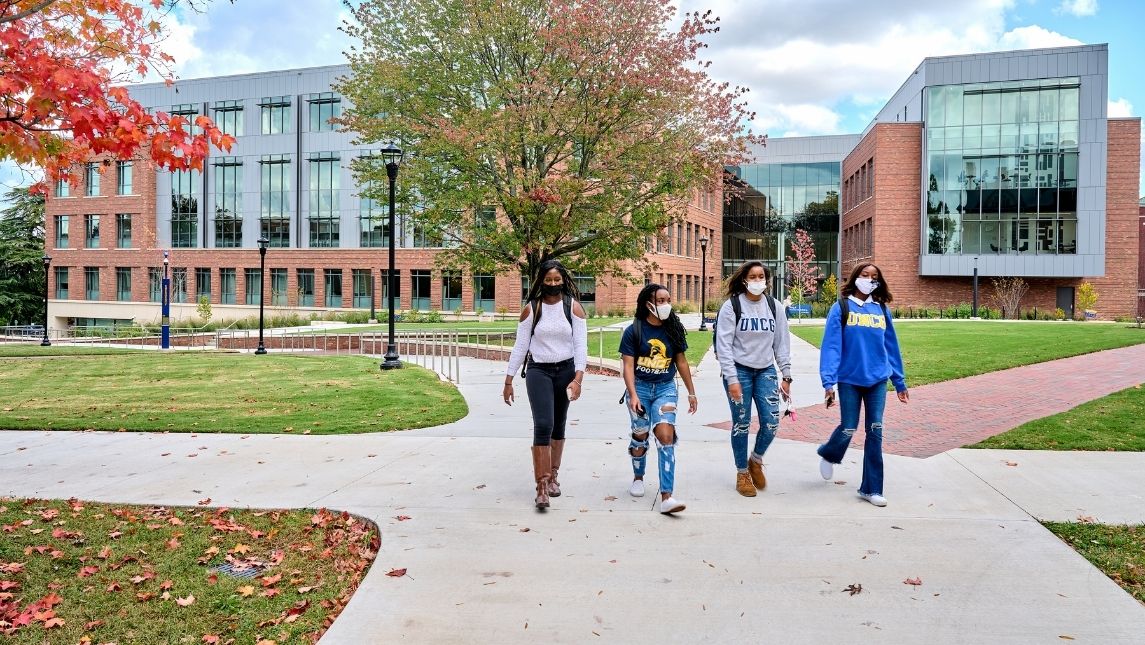 four students walk on a sidewalk from the Nursing Instructional Building among fall trees