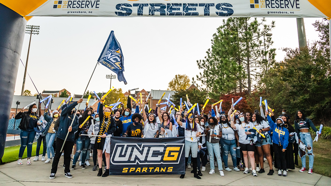 A crowd of students decked out in blue and gold UNCG gear holding LED blue and gold batons stand and cheer behind a UNCG Spartans banner and a UNCG Spartans flag