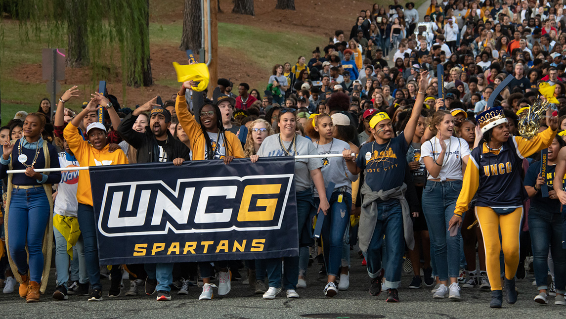 Crowd of students in blue and gold marching down the street holding a UNCG Spartans banner