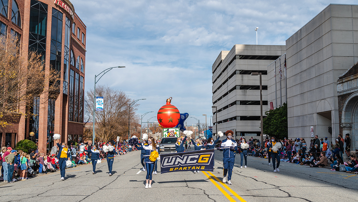 UNCG cheerleaders hold a UNCG Spartans banner and wave at the crowd while marching down the street in Downtown Greensboro as part of the Holiday Parade