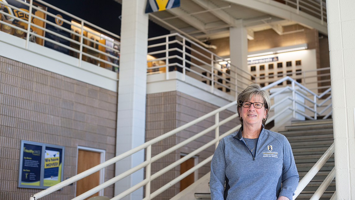 Researcher in UNCG sweater standing in the Coleman building with a staircase behind her.
