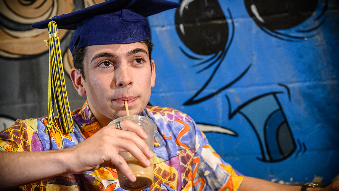 Colby Borges drinking iced coffee in a graduation cap at Common Grounds