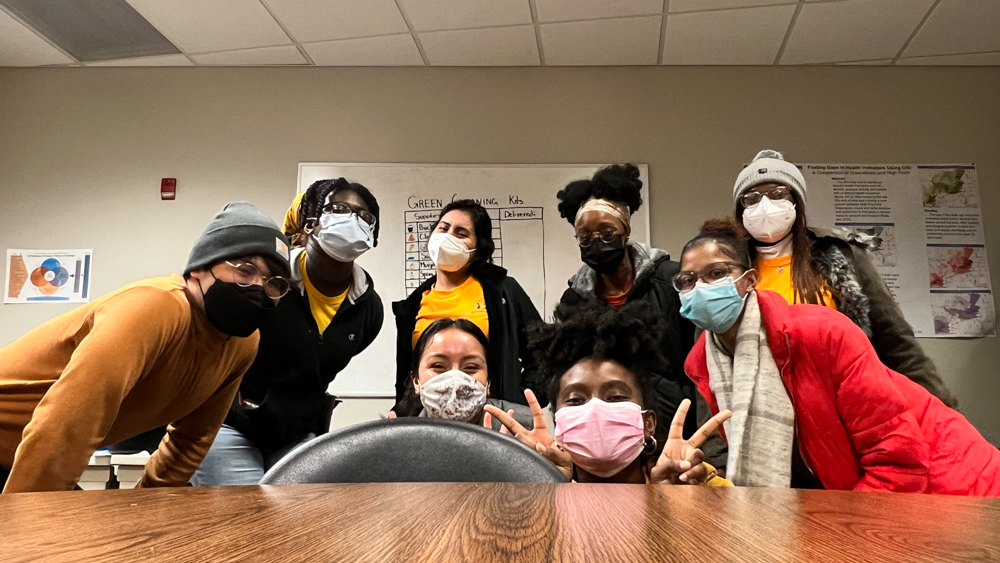 A group of students wearing masks smile in front of a dry erase board.