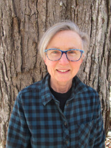 A woman wearing glasses stands in front of a tree.