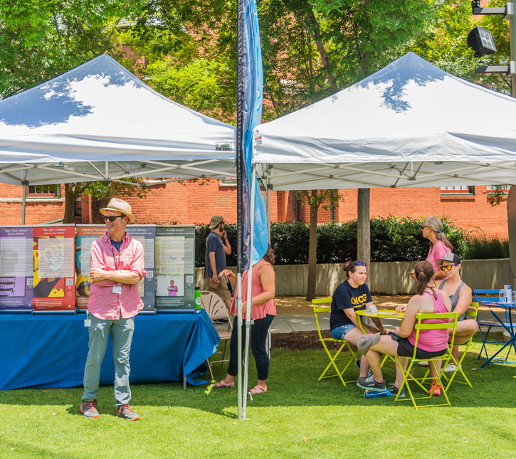 people having a conversation at a table at a park, a man standing at a display table