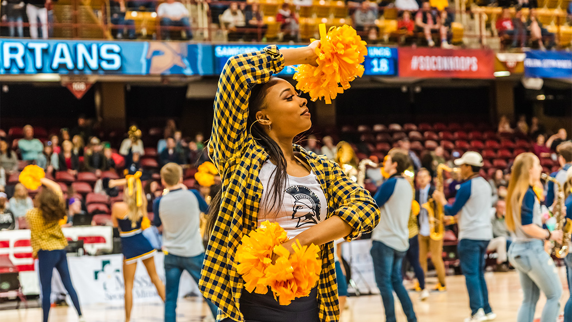 Cheerleader in a checkered shirt holds up pompoms.