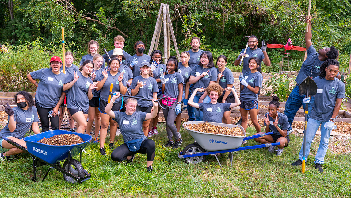 Large crowd of students gather around spades and wheelbarrows in garden.