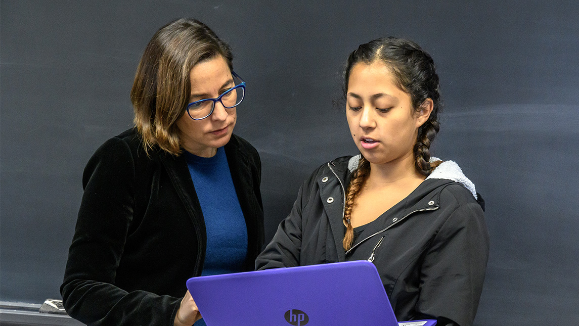 Dr. Cabello Hutt stands in front of chalkboard while student shows her a laptop.