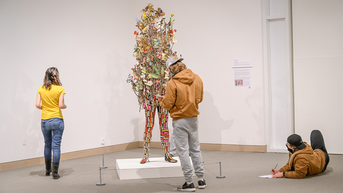 Three students stand or sit around a museum exhibit made of cloth birds and flowers on a mannequin.