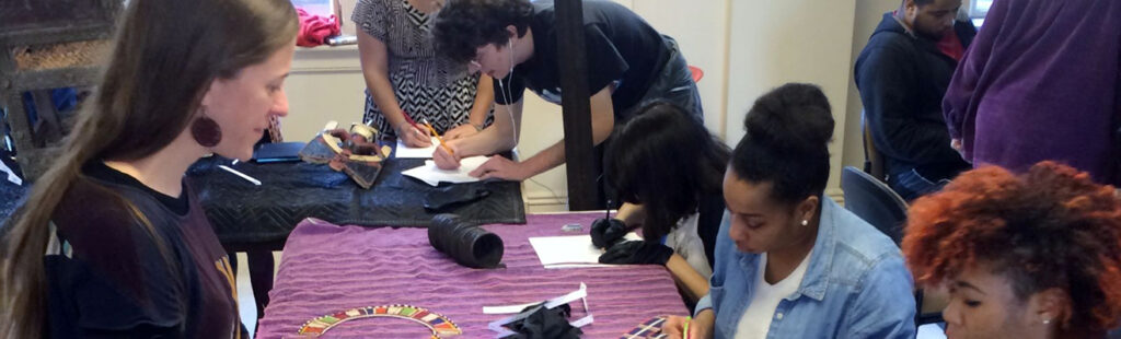 An Art History Professor stands before two students seated at a table as other students mill about in the background.