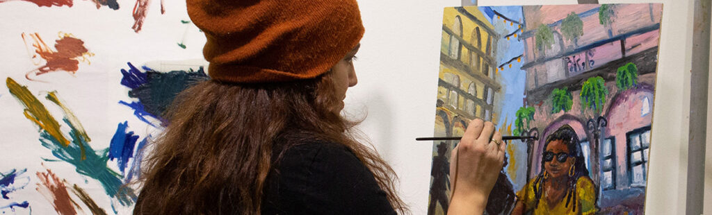 A student wearing an orange beanie paints at an easel.