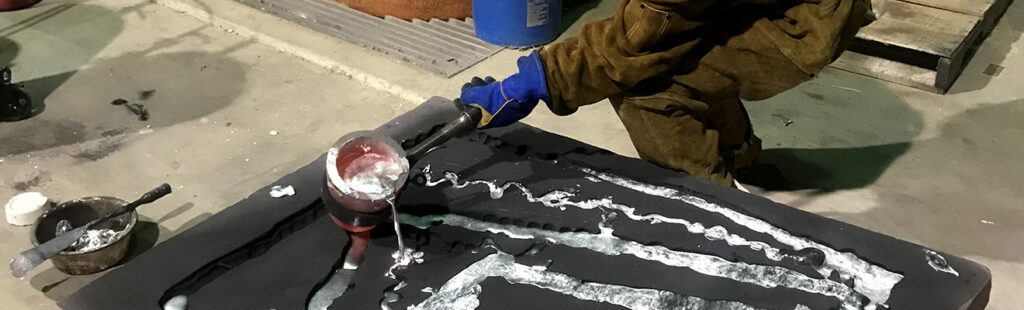 A student wearing a yellow hardhat and face shield pours molten metal into a mold.