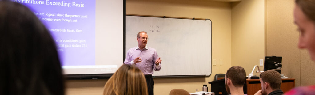 A UNCG Bryan Accounting (BS) professor addresses a classroom of business students. He is in the background of the shot, with some of the shot filled with the backs of students heads. He is standing in front of a projector screen, wearing a purple button down shirt.