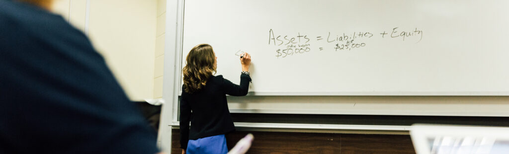 A UNCG Bryan School professor is standing facing a white board in a classroom. She is writing an equation.She is wearing a blue dress and a black jacket.