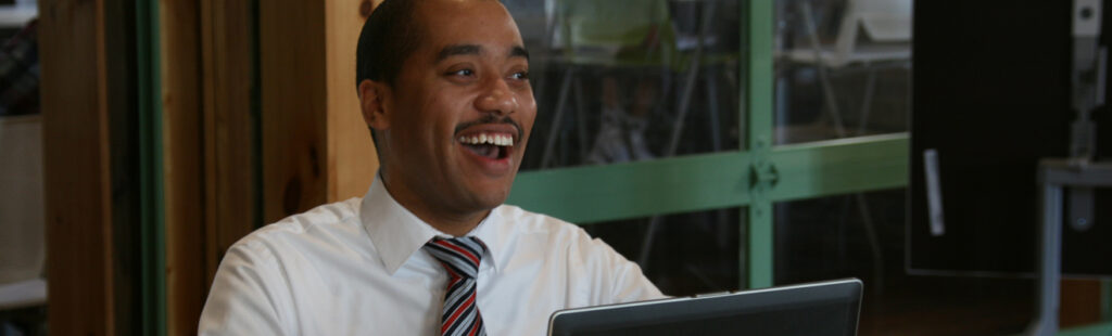 A UNCG Bryan School student is sitting in a room at his laptop. He is smiling and laughing. He has a moustache and is wearing a tie.