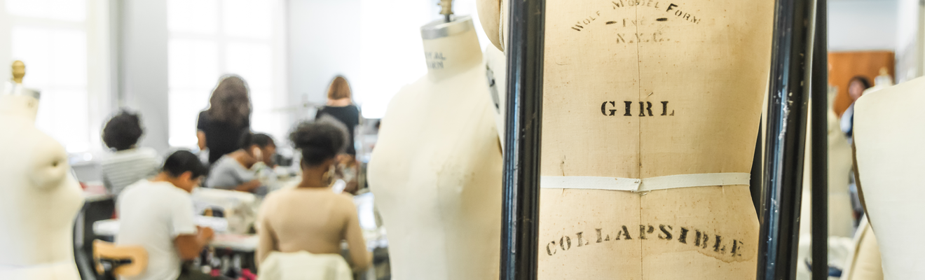 UNCG Bryan students sit at sewing machines. They are blurred and in the background of the photo to the left of the image. In the foreground to the right are two mannequins, not dressed in any fabrics.