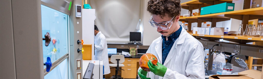 Male student wearing goggles and lab coat looks at orange substance in test tube