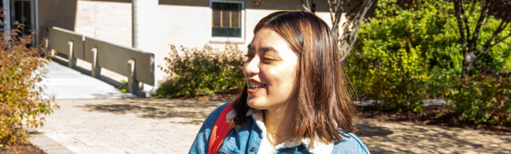 A UNCG student stands outside of the Bryan School. She is smiling and wearing a jean jacket, looking off camera to the left of the photo.