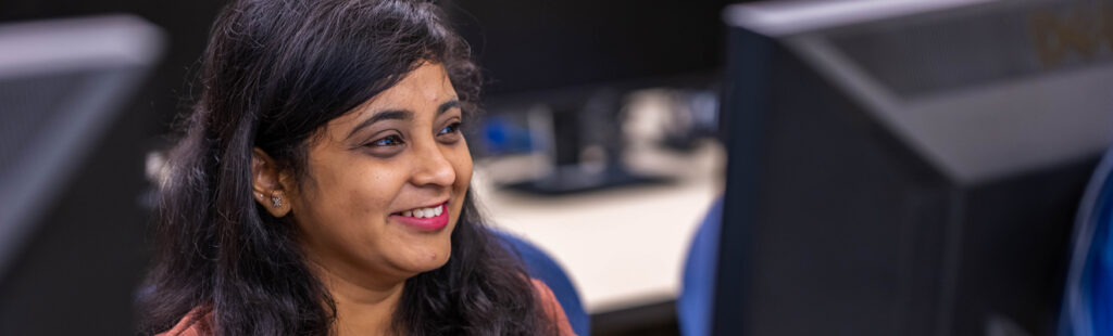 A UNCG student sits amongst computers on campus. The screens are facing away from the camera. She is smiling, looking at her screen and focusing on her work.