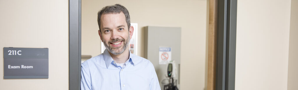 A UNCG Bryan Economics professor stands in the doorway of an exam room, smiling and facing the camera. He is wearing a blue button down shirt.