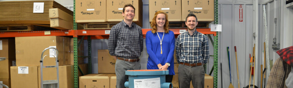 Three UNCG Supply Chain Management certificate students stand in front of a stack of boxes in a warehouse. They are shoulder-to-shoulder, smiling for the camera, with a dolly to their right.