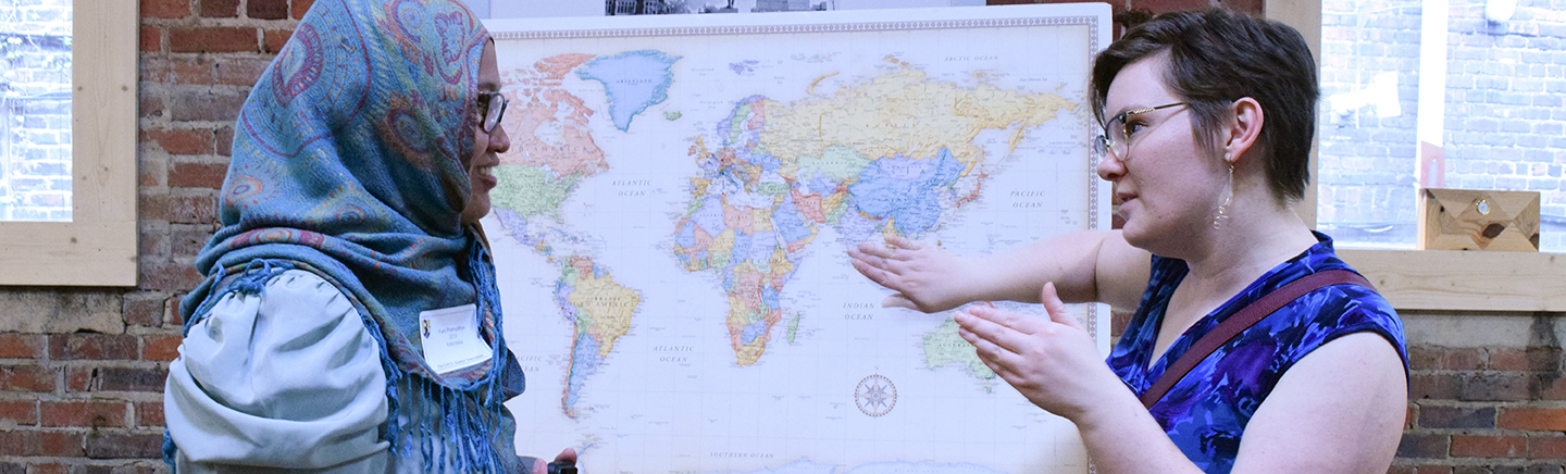 A female professor discusses the world map on a tripod stand and points out locations to a female student