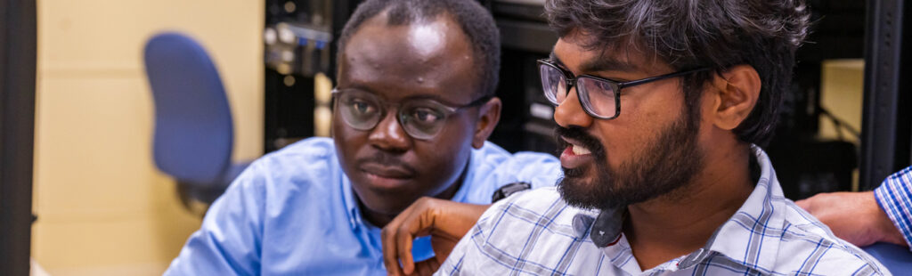 UNCG students sit in a computer lab in the middle of a discussion. They are looking to the left of the photo. Both are wearing button down shirts and glasses.