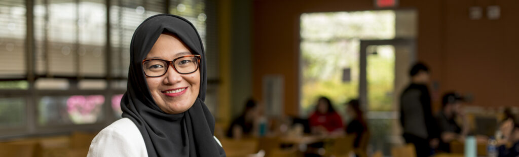 A professor is standing among his students in a UNCG Bryan Master of Science in International Business course. Some are turned back to see him chat with a student. The professor, who is wearing glasses, is smiling while speaking to the student, who is in the lower left corner of the image.