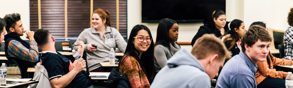 This is a wide shot of a UNCG Bryan School course. A female student with glasses sits in the middle of the room, looking at her camera to the right and smiling.