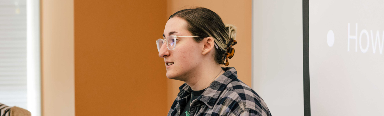 UNCG Bryan MS inUNCG Bryan MS in IT and Management student stands and presents during class. She is wearing a black and gray plaid shirt and glasses.