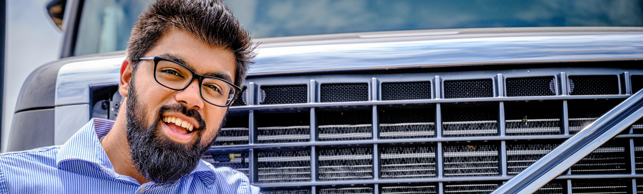 UNCG Bryan MS in IT and Management student stands next to a truck during a photoshot at an internship. He is smiling and leaning on the grill of the semi