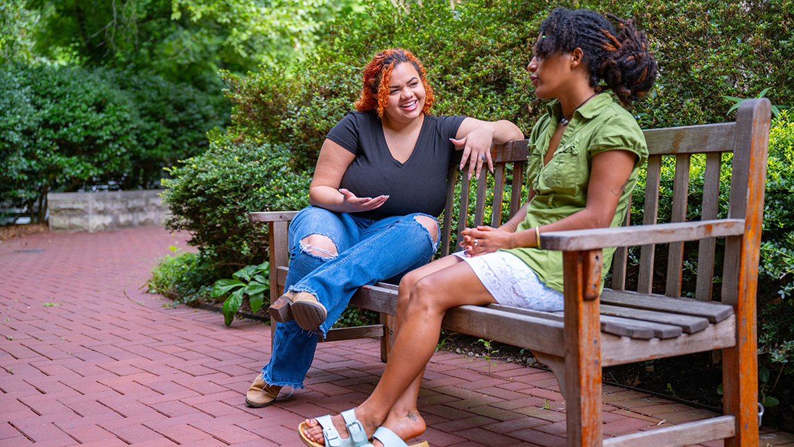 Two women speak with one another on a bench with trees behind them.