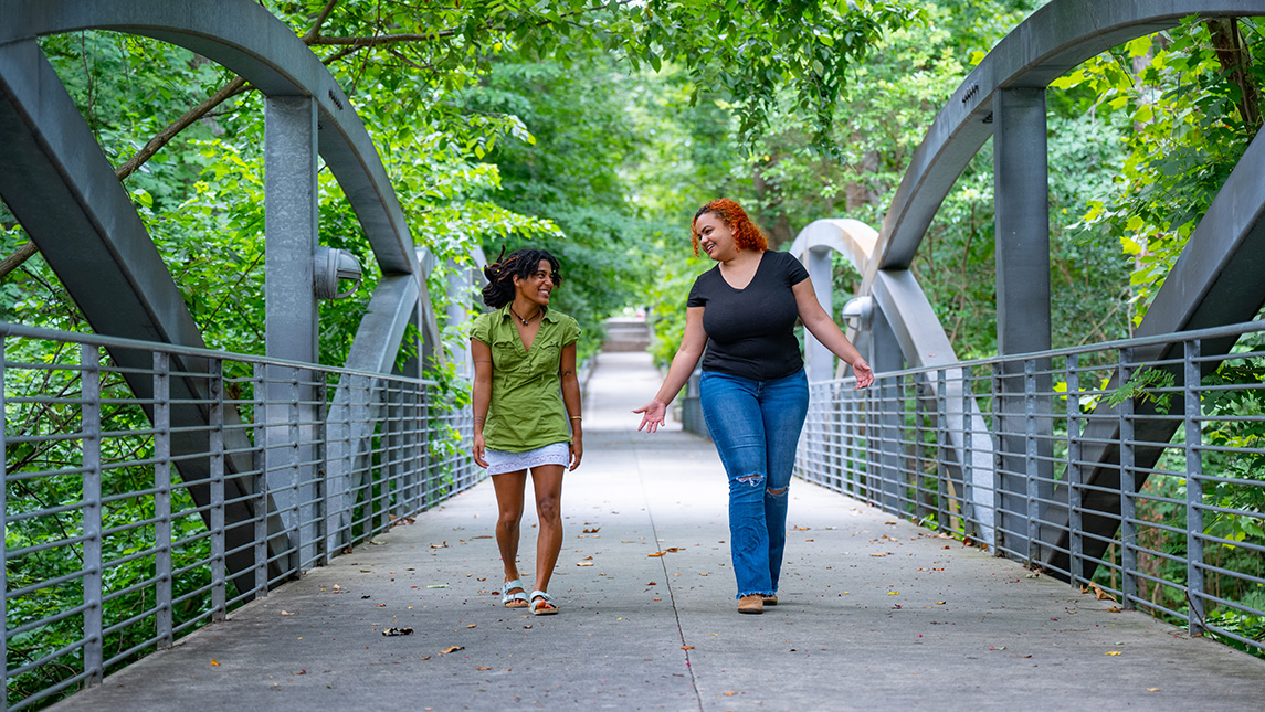 Two women walk side-by-side over a bridge with greenery around them.