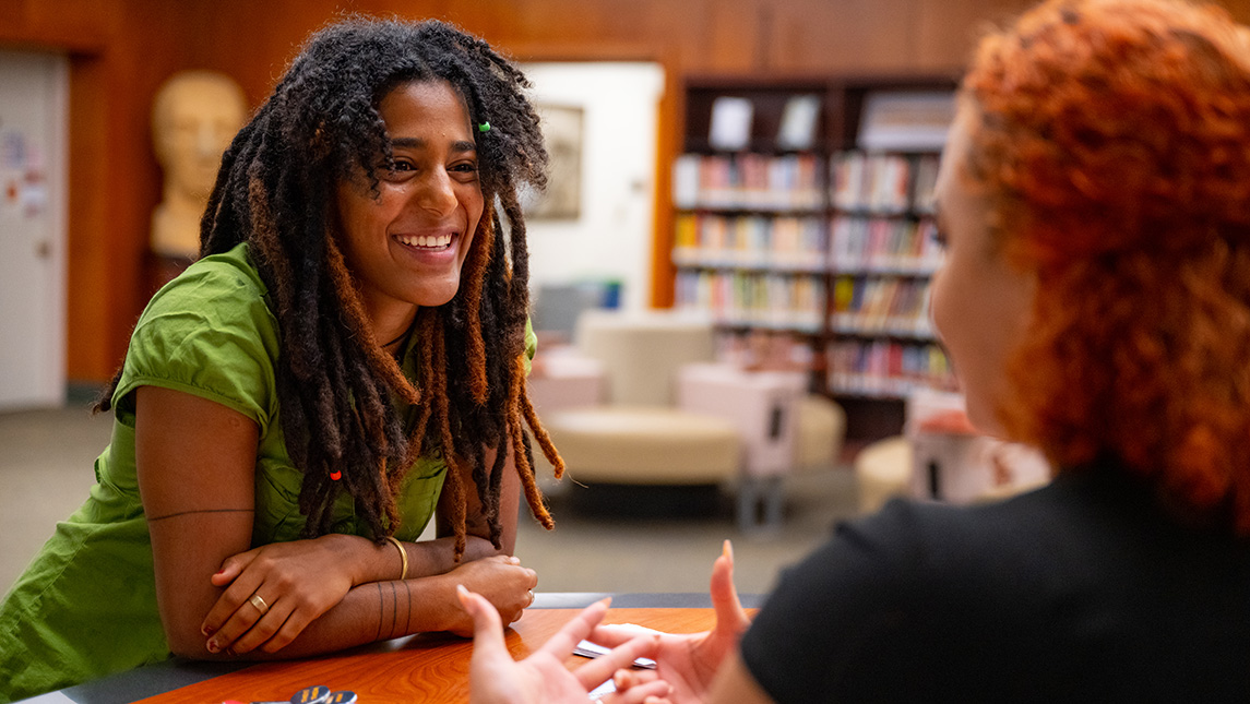 Two woman speak with one another in a library with books behind them.