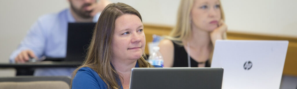Three UNCG Bryan PhD in Business Administration - Management students are sitting in a classroom, all with laptops open in front of them, taking in a lecture. Two are blurry in the background. In focus to the center left of the photo is a female student wearing a blue shirt.