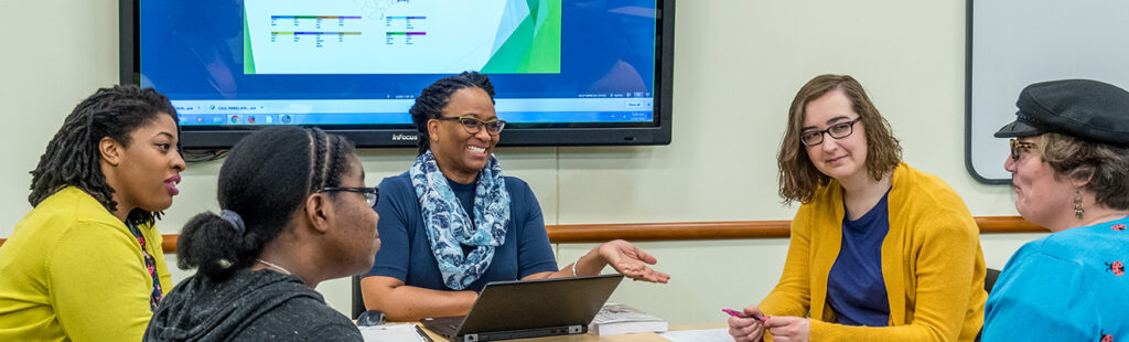 Library and Information Science students conversing in a classroom setting with a powerpoint presentation pulled up on the screen behind them