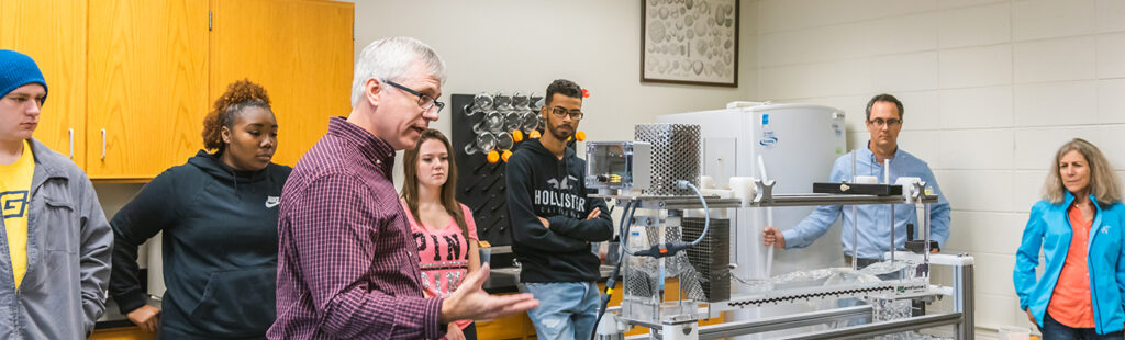 Geography professor standing in middle of science classroom teaches Geography degree students about a medium-sized geography apparatus on the table